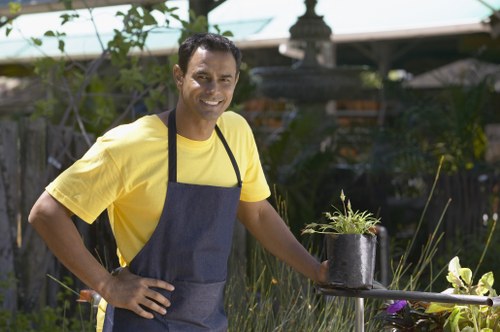 A man with medium skin tone, short dark hair, and a friendly smile is standing outdoors in a garden during daylight, wearing a bright yellow T-shirt and a dark grey apron. He is holding a small potted plant with green foliage and some purple flowers in his right hand, while his left hand rests on his hip. The garden features a well-maintained lawn with dense, lush grass, bordered by various shrubs and leafy plants, some flowering, and a weathered wooden fence in the background. A decorative stone lantern or garden ornament is visible behind him, along with garden tools or equipment partially obscured by foliage. The environment appears sunny with natural light illuminating the scene, suggesting a spring or summer setting. This image, associated with gardening services like hedge trimming and outdoor maintenance, highlights the care and attention involved in garden landscaping in Charlton, Kent, reflecting the local outdoor gardening expertise of Hedge Trimming Charlton.