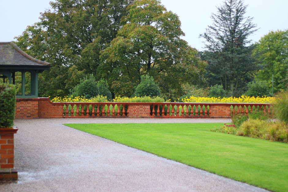 A wooden picket fence with weathered, dark-stained vertical slats runs along the edge of a small fenced garden. The slats are evenly spaced and topped with pointed tips. Behind the fence, a neatly mai